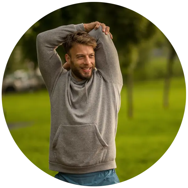 Man stretching outdoors in a grey hoodie, smiling, in a green park setting, promoting wellbeing and self-care during winter months.