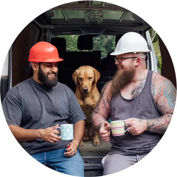 Two construction workers enjoying coffee breaks in a van, one wearing a red hard hat and the other a white hard hat, with a golden retriever sitting between them.